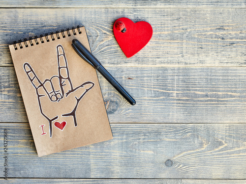 Notebook of Kraft paper, which is painted with an old black marker, the symbol of ASL (American Sign Language) ILY and a wooden red heart with a ladybug