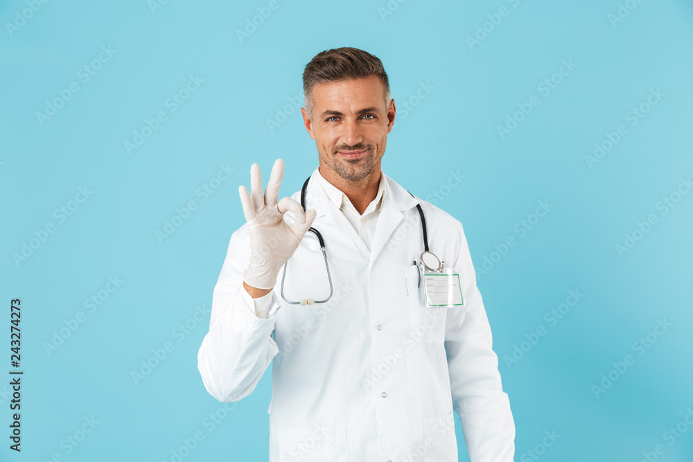 Handsome mature man doctor posing isolated over blue wall background showing okay gesture. Handsome mature man doctor posing isolated over blue wall background showing okay gesture.