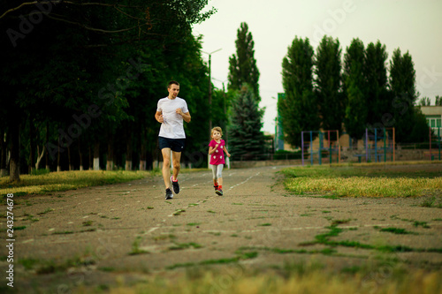 Fototapeta Naklejka Na Ścianę i Meble -  Athletic young father and little daughter running in stadium at sunset.