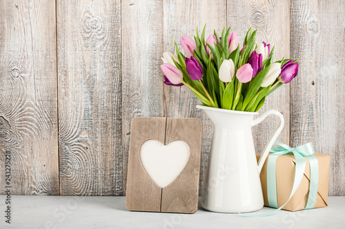 Fresh colorful tulips in a jug, gift box and heart shaped frame on table