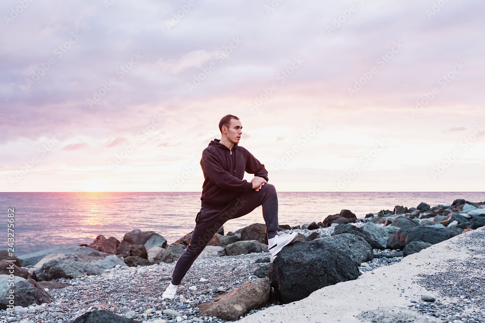 Side view of a slim tall guy athlete doing a warm-up on a rocky seashore against the backdrop of the sunset. Concept of sports training after a hard day. Copyspace Side view of a slim tall guy athlete doing a warm-up on a rocky seashore against the backdrop of the sunset. Concept of sports training after a hard day. Copyspace