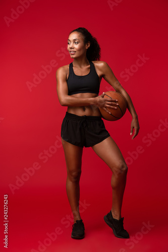 African sports fitness woman basketball player posing isolated over red wall background holding ball. © Drobot Dean