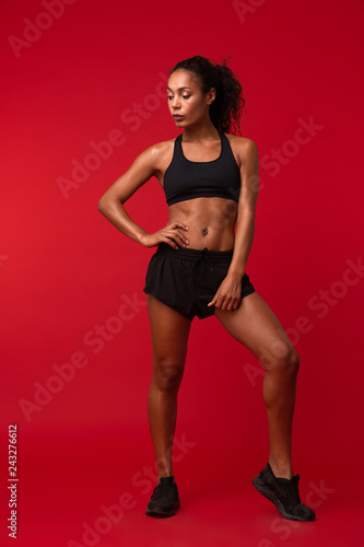 Portrait of pretty african american woman in black sportswear standing, isolated over red background © Drobot Dean