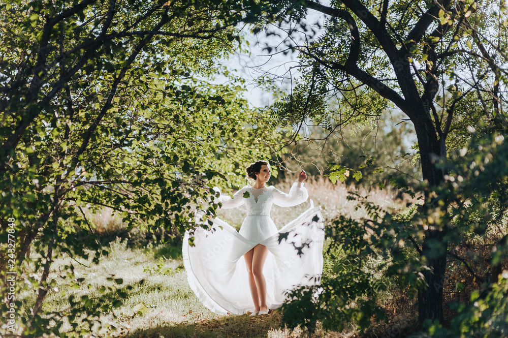 Beautiful bride in lace dress is dancing on the background of nature and green trees. Portrait of a young bride in backlight. Wedding photography. Cheerful girl.