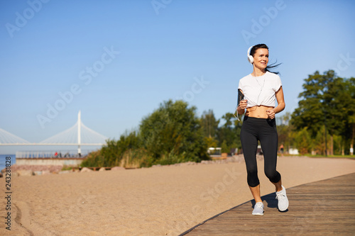 Content pretty young lady in headphones wearing smartphone armband looking around while running over path on summer beach © pressmaster