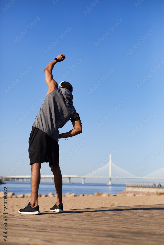 Rear view of black man in sportswear listening to music via headphones and performing side bend stretch while contemplating beach in morning