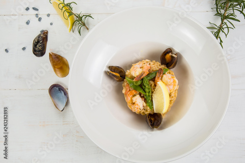 An overhead photo of a seafood risotto on teal textures, with a fork and a spoon, a glass of white wine, and a place for text