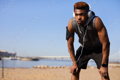 Serious tired young African man in wired headphones being without strength leaning on knees and trying to catch breath after running on beach