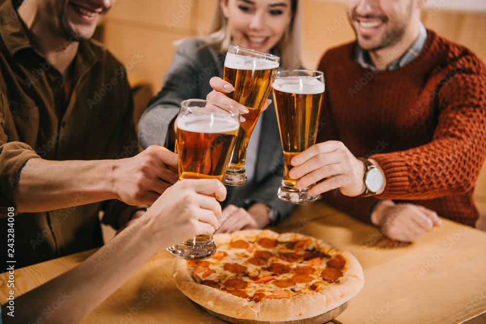 cropped view of friends clinking beer near pizza in bar