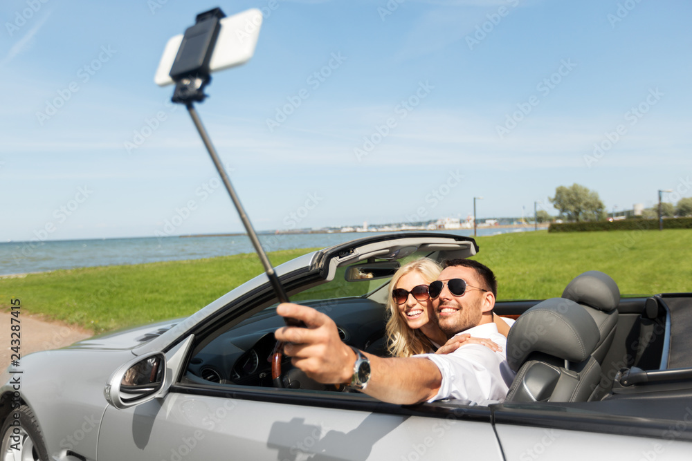 road trip, leisure, technology and people concept - happy couple in convertible car taking picture by smartphone on selfie stick