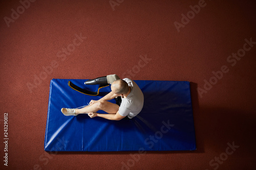 Overview of young paralympic competitor with artificial right leg sitting on blue mat and tying shoelace on left sneaker photo