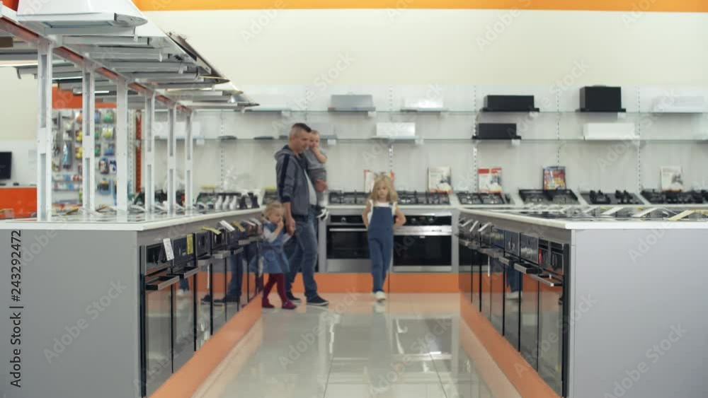 Father walking with two little daughters and baby boy on arms through aisle in home appliance store. Family talking and looking at kitchen stoves Father walking with two little daughters and baby boy on arms through aisle in home appliance store. Family talking and looking at kitchen stoves