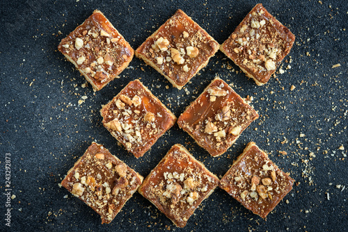 Homemade caramel shortbread squares cookies with nuts laid in the shape of a square on black background with crumbs.