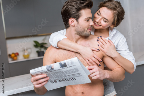 happy woman hugging handsome boyfriend with business newspaper in kitchen