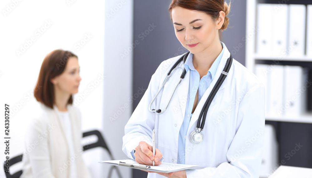 Doctor woman at work. Portrait of female physician filling up medical form while standing near reception desk at clinic or emergency hospital. Patient woman sitting at the background. Medicine concept Doctor woman at work. Portrait of female physician filling up medical form while standing near reception desk at clinic or emergency hospital. Patient woman sitting at the background. Medicine concept