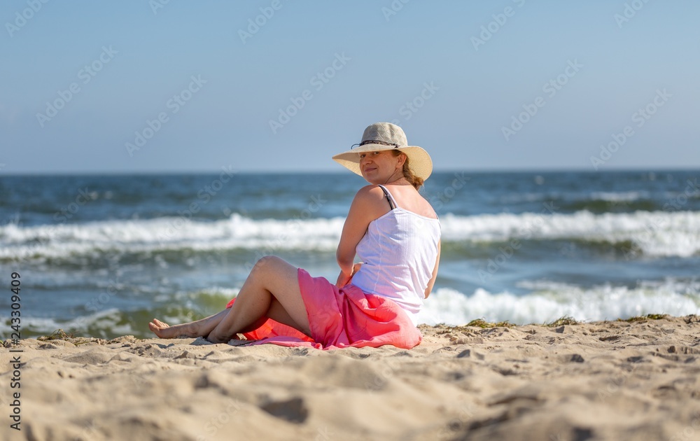 Beautiful blonde woman in hat and skirt sitting on sea shore.