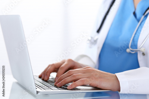 Doctor typing on laptop computer while sitting at the glass desk in hospital office. Physician at work. Medicine and healthcare concept