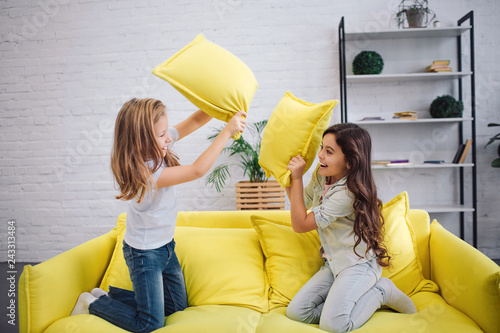 Two cheerful teenagers have pillow fight on sofa in room. They stand on knees in ront of each other. photo