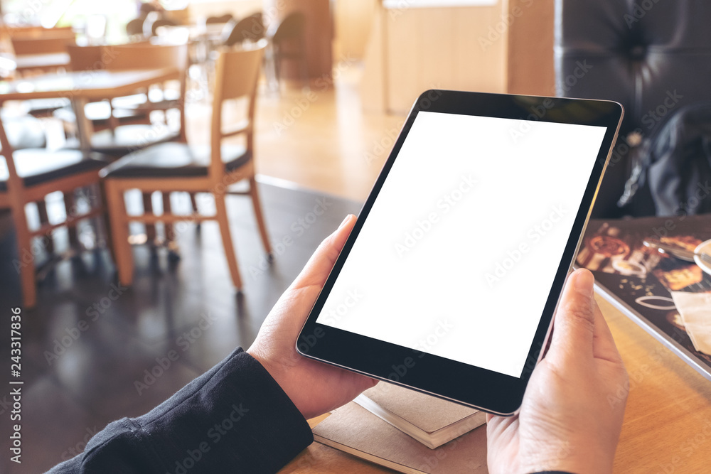 Mockup image of hands holding black tablet pc with blank screen with notebook and bread on wooden table in cafe