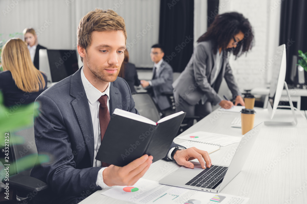 focused young businessman holding notepad and using laptop in open space office focused young businessman holding notepad and using laptop in open space office