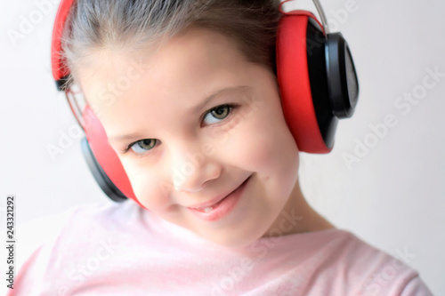Portrait of caucasian white pretty little girl listening to music with red headphones on beige neutral background. Beautiful brunette smiling green eyed girl listen music with big earphones.