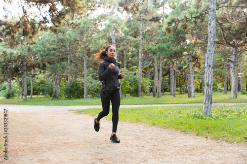 Fototapeta Naklejka Na Ścianę i Meble -  Image of sportswoman 20s wearing black tracksuit working out, while running through green park