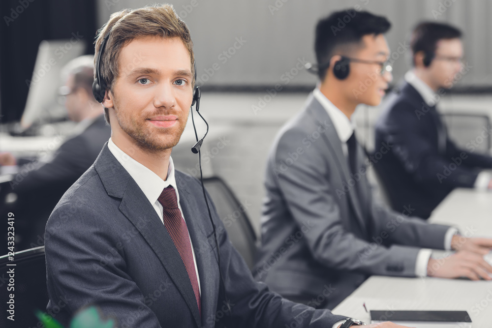 handsome young businessman in headset smiling at camera while working with colleagues in office handsome young businessman in headset smiling at camera while working with colleagues in office