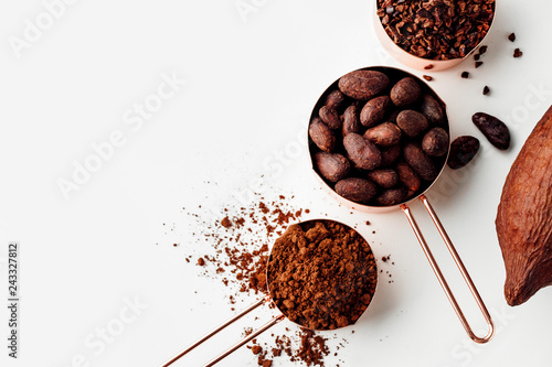 Rose gold measuring cups of cocoa beans, cacao nips, cocoa powder and cocoa pods on a white background, flat lay healthy food concept