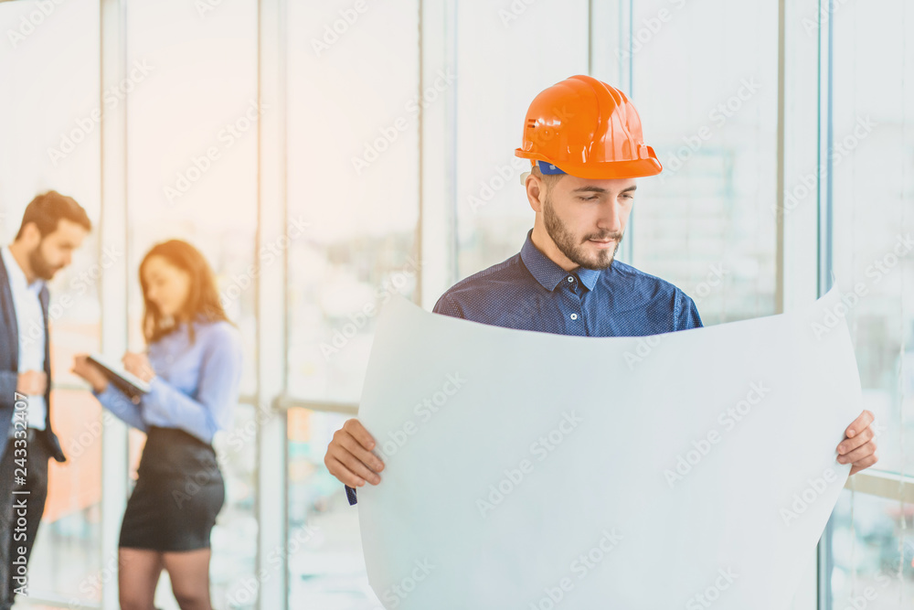 The architect is in a cask of orange color. In the office against the background of their colleagues.