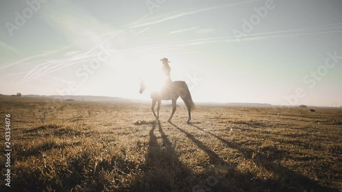 Cowgirl riding brown horse across empty field at sunrise