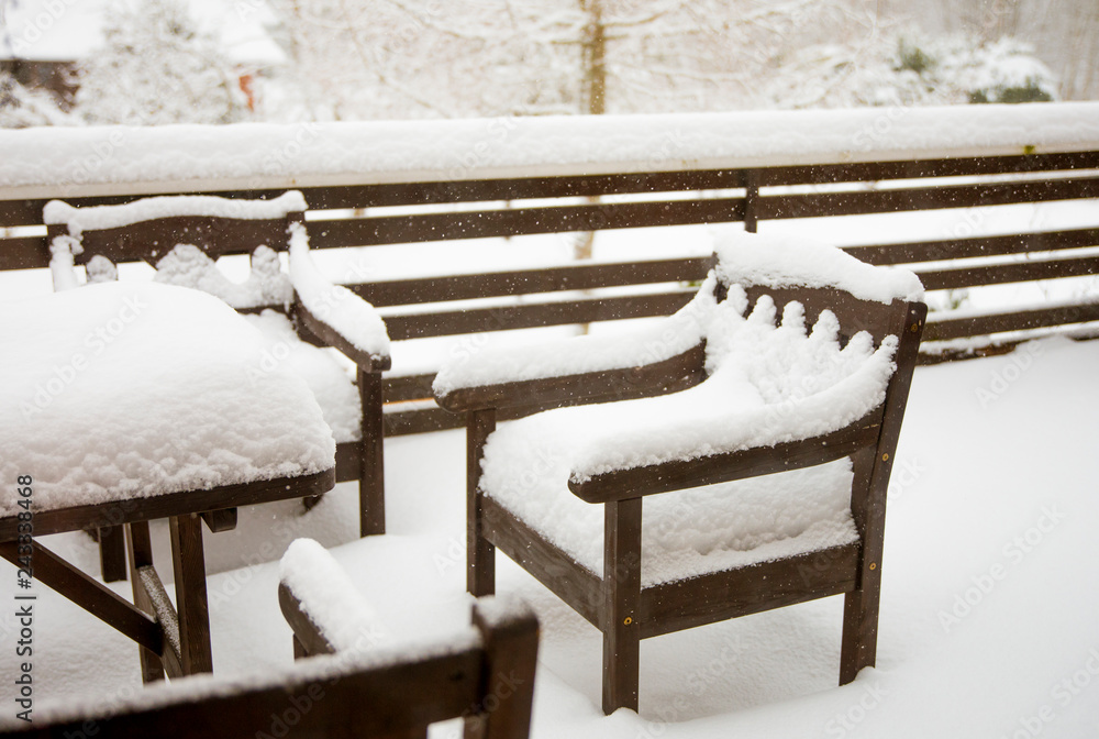 Fresh clean snow covering brown wooden Mahogany color stained garden chairs and table on home balcony. Garden furniture maintenance after cold winter concept.