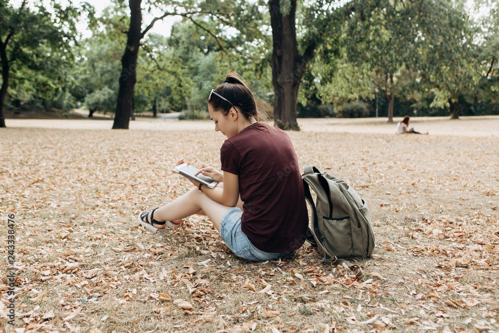 A young girl with a backpack sits in the autumn park and uses the tablet. She watches a video or reads or online or something else.