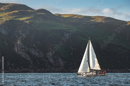 Touristic yacht sailing next to the Northern Ireland steep rocks. Green covered heels, shoreline. Outdoor activity. Perfect background for the creating of various kinds of collages and illustrations.