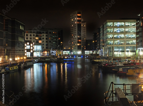 clarence dock in leeds at night with brightly illuminated buildings reflected in the water and boats moored along the sides