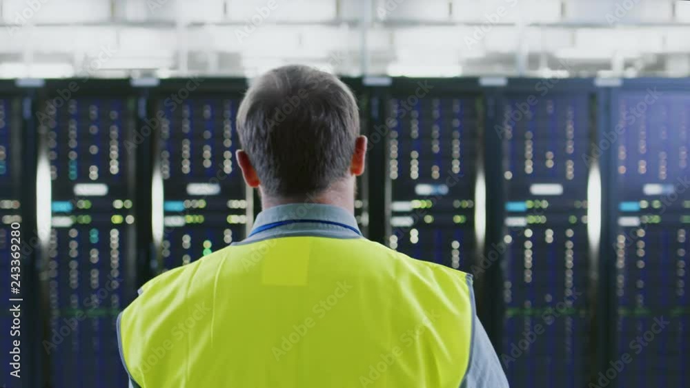 Follow Back Shot of an IT Administrator in High Visibility Vest Walking Towards a Server Rack in Data Center Room. Follow Back Shot of an IT Administrator in High Visibility Vest Walking Towards a Server Rack in Data Center Room.