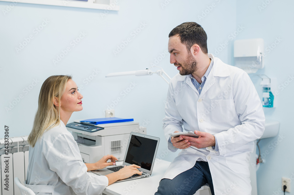 Two doctors colleagues are sitting at the work place and using modern technology for their work, in white coats, focused Two doctors colleagues are sitting at the work place and using modern technology for their work, in white coats, focused