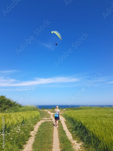 Woman watching adult paraglider near cliff along baltic sea coastline and green meadow wheat field at Boltenhagen Coast, germany, for themes of extreme sports, lifestyle, escapism