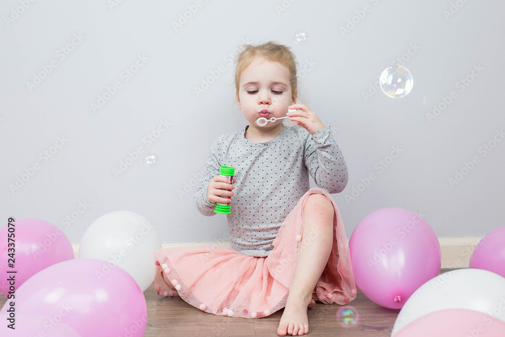 Beautiful little blonde girl, has happy fun cheerful smiling face, pink dress, soap bubble blower. Portrait with pink and white balloons