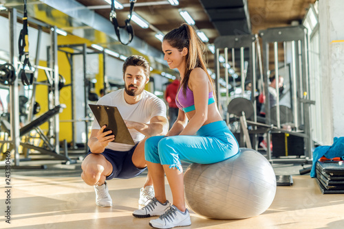 Smiling Caucasian sporty woman sitting on pilates ball and looking at results of training that her personal trainer showing her. Trainer crouching next to her.