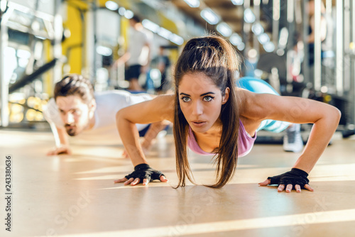 Close up of beautiful Caucasian woman doing push-ups in gym. In background her personal trainer showing exercises.