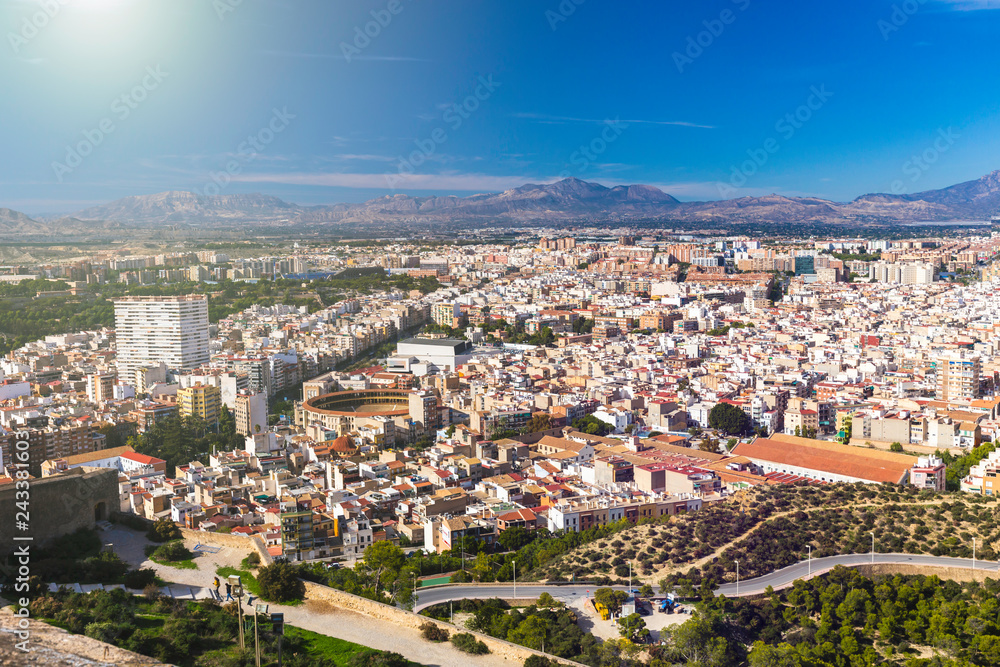 Panoramic view of the city of Alicante, Spain. Block of apartment buildings, parks, roads, houses, palm trees and bullring against the background of a mountain landscape in the blue sky