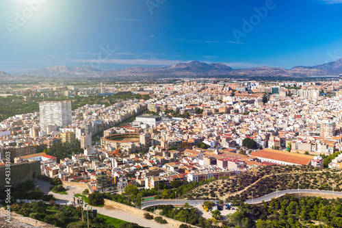 Fototapeta Naklejka Na Ścianę i Meble -  Panoramic view of the city of Alicante, Spain. Block of apartment buildings, parks, roads, houses, palm trees and bullring against the background of a mountain landscape in the blue sky