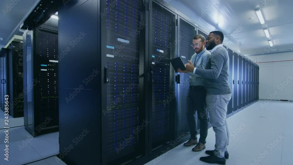 Bearded IT Technician in Glasses with a Laptop Computer and Black Male Engineer Colleague are Talking in Data Center while Working Next to Server Racks. Running Diagnostics or Doing Maintenance Work.