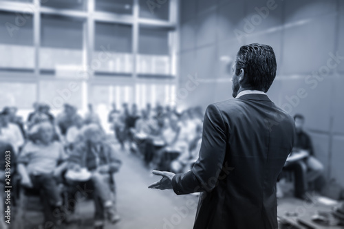 Rear view of speaker giving a talk at business meeting. Audience in the conference hall. Business and Entrepreneurship concept. Black and white blue toned image.