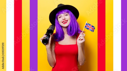 Portrait of young style hipster girl with purple hairstyle with binoculars and Great Britain flag in hand on yellow background