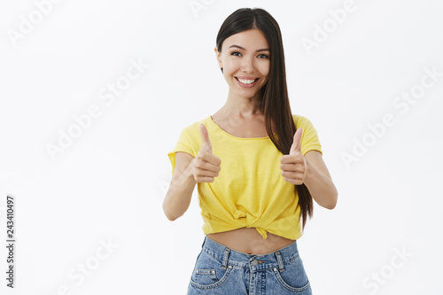 Friendly attractive and outgoing supportive tanned female with acne and natural beauty without makeup showing thumbs up and smiling broadly approving and liking awesome idea against grey wall