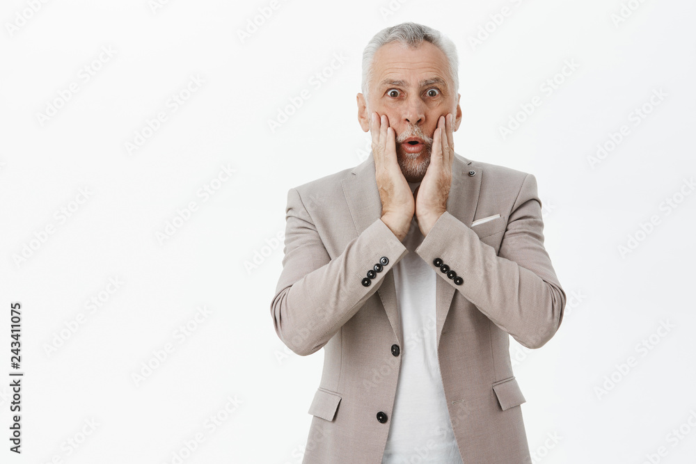 Portrait of surprised and impressed grandfather with white beard and hair gasping touching face from amazement and shock staring astonished at camera reacting to unexperted news over grey wall
