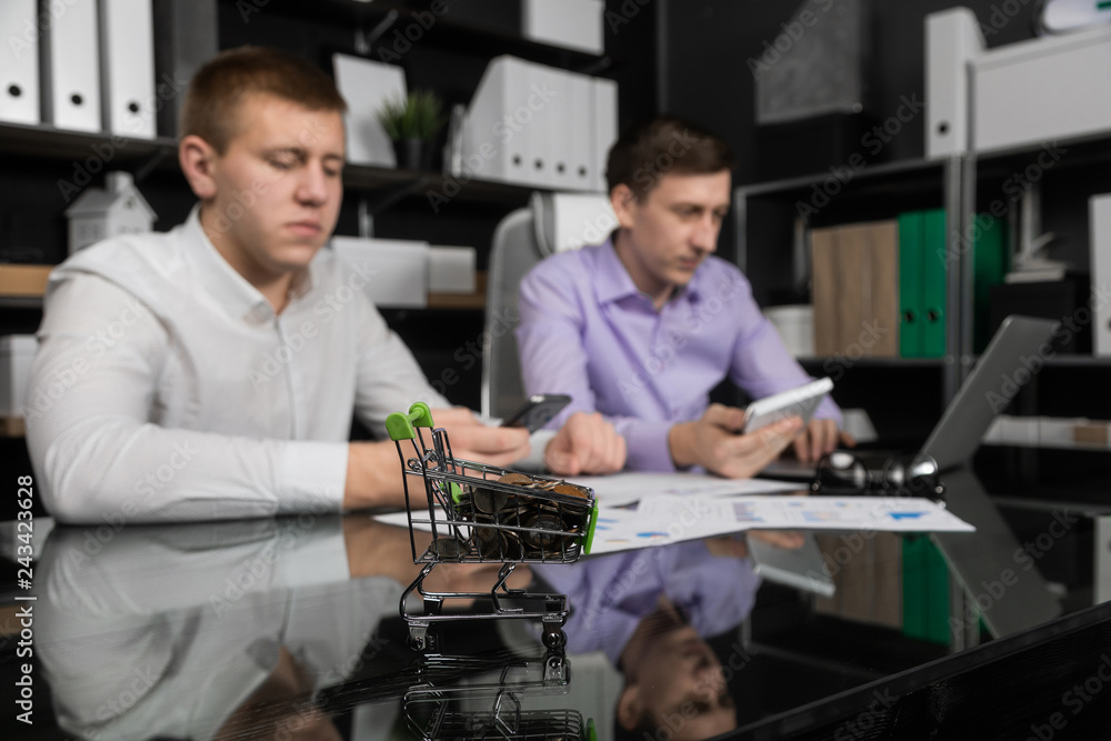 Dummy shopping cart with coins on the office table on the background of two businessmen