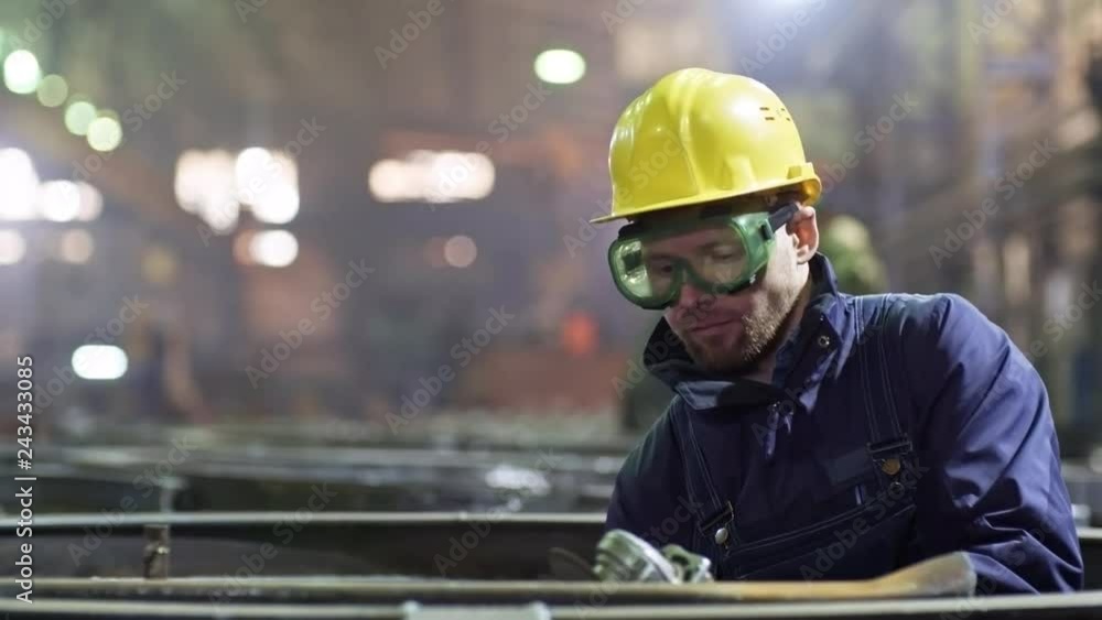 Tracking shot of young male worker in hard hat and safety goggles using angle grinder machine on steel part at metal fabrication plant Tracking shot of young male worker in hard hat and safety goggles using angle grinder machine on steel part at metal fabrication plant