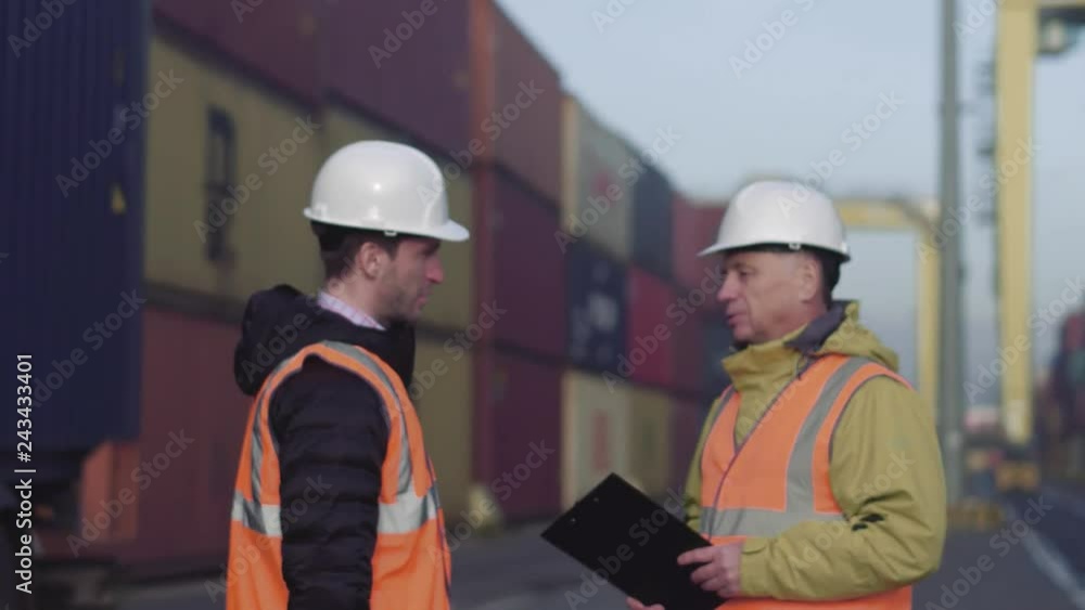 Port manager and a colleague tracking inventory while standing together by freight containers on a large commercial shipping dock Port manager and a colleague tracking inventory while standing together by freight containers on a large commercial shipping dock